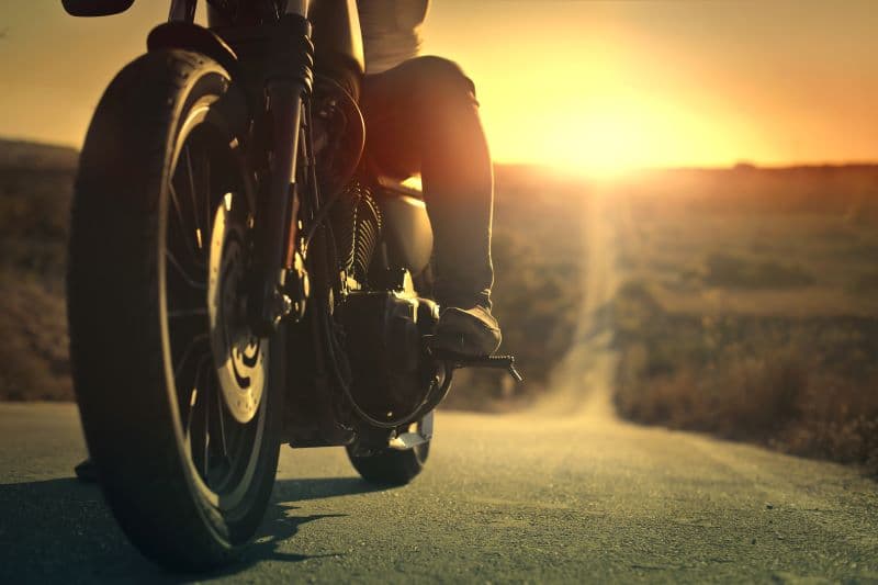 Moto Harley-Davidson con pilota, dettaglio ruota anteriore e motore, al tramonto su strada. Invita a un test ride.