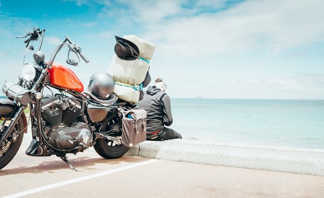 Harley-Davidson rossa con bagagli e casco, parcheggiata in riva al mare. Il motociclista in abbigliamento tecnico ammira il panorama.