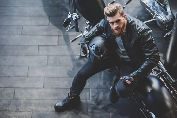 Uomo con barba e giacca in pelle nera seduto su moto Harley-Davidson, tiene casco. Incarna lo stile e la passione per i motori.
