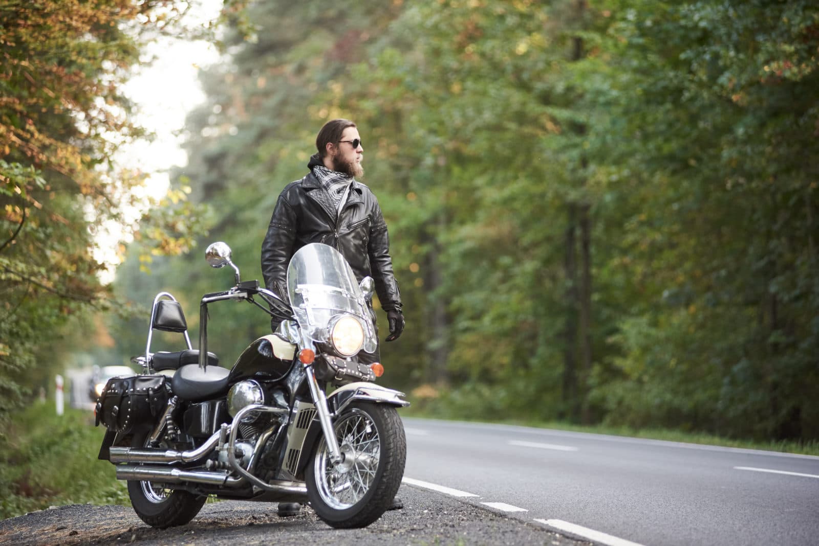 Motociclista con giacca in pelle e occhiali da sole, in posa accanto a una moto cruiser Harley-Davidson su strada. Stile e abbigliamento icon