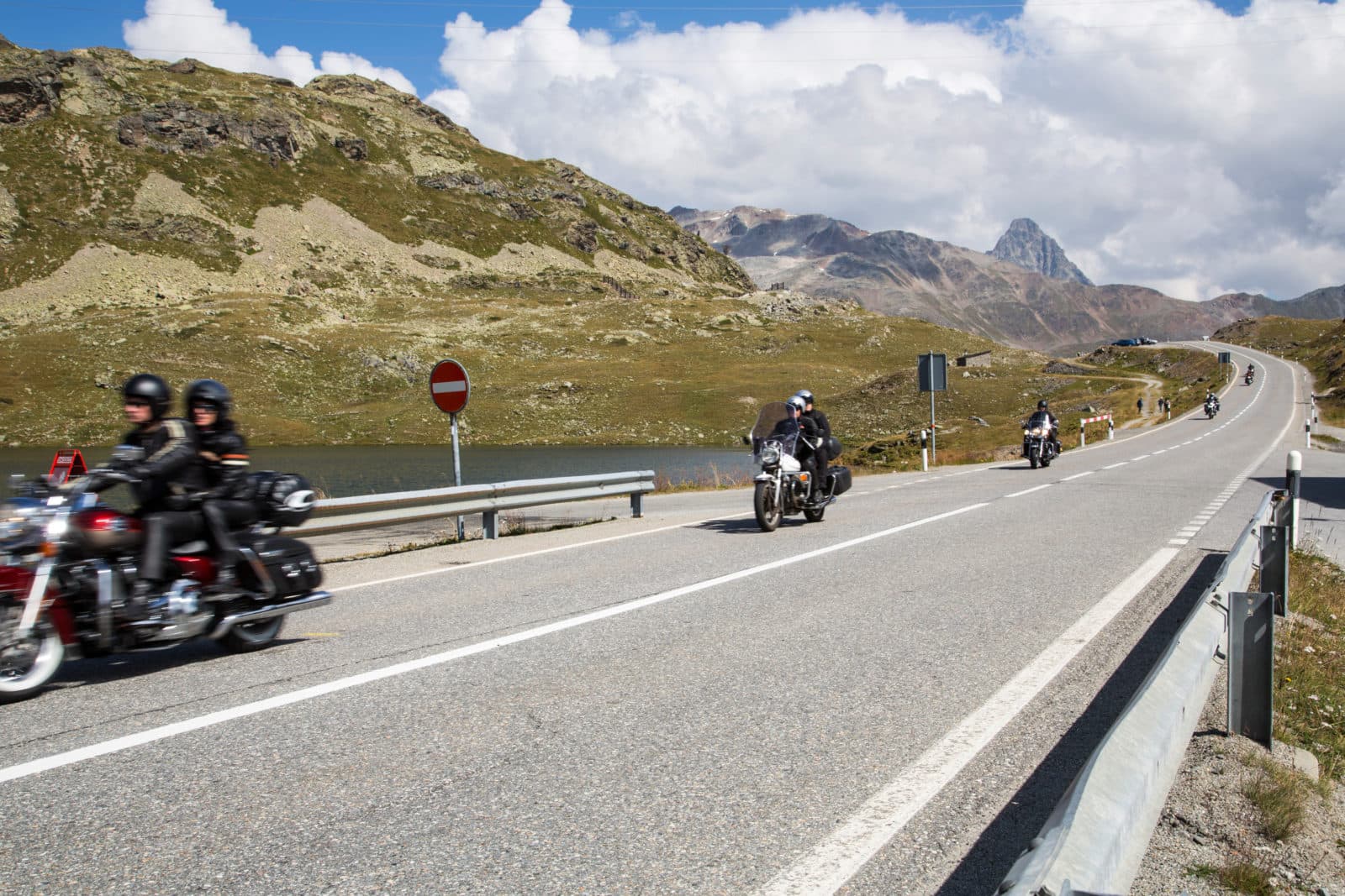 Gruppo di motociclisti in sella a moto, inclusa una Harley-Davidson, su una strada di montagna panoramica. Indossano abbigliamento