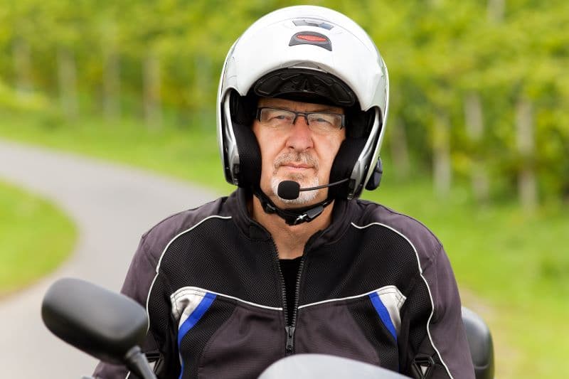 Motociclista con casco modulare argento, occhiali e giacca tecnica, dotato di interfono, in sella alla sua moto, pronto per un test ride.