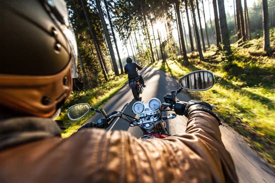 Esperienza di guida in moto: vista in prima persona di un motociclista con abbigliamento tecnico, seguendo un'altra moto su strada forestale illuminata dal sole.
