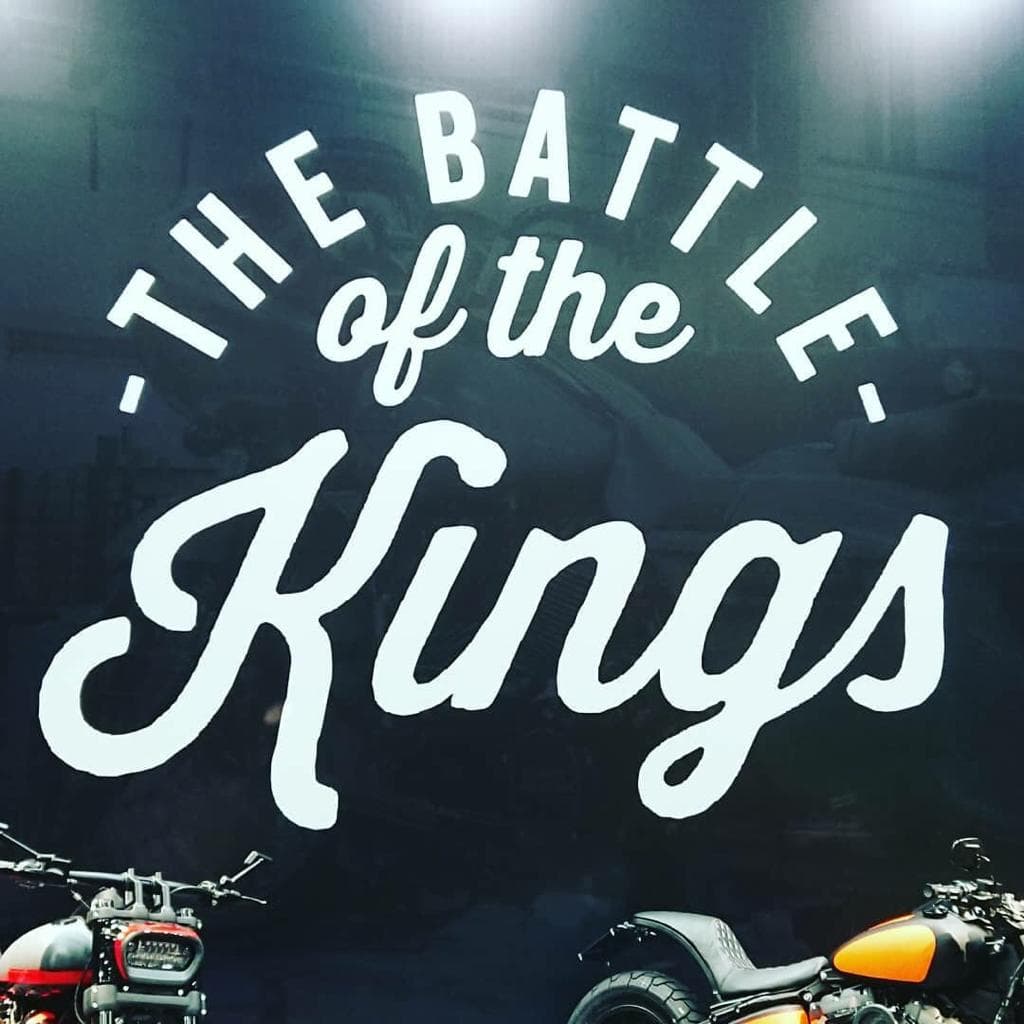 Insegna promozionale Harley-Davidson 'The Battle of the Kings' con lettering bianco e due moto custom in primo piano, una rossa e una arancione.