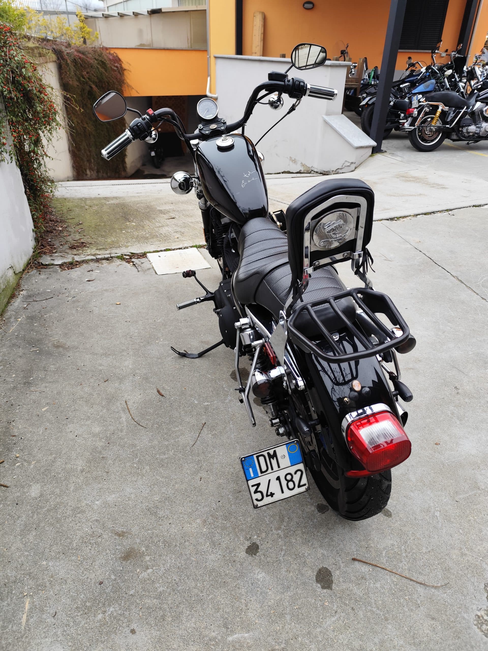 Harley-Davidson nera personalizzata con manubrio ape hanger, sissy bar e portapacchi, parcheggiata su cemento. Targa italiana DM 34182.