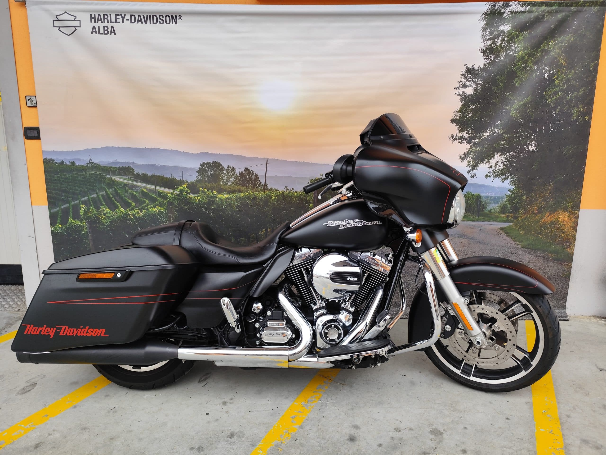 Moto Harley-Davidson touring nera opaca, motore 103, con dettagli rossi, esposta davanti al banner Harley-Davidson Alba con vigneti al tramonto.