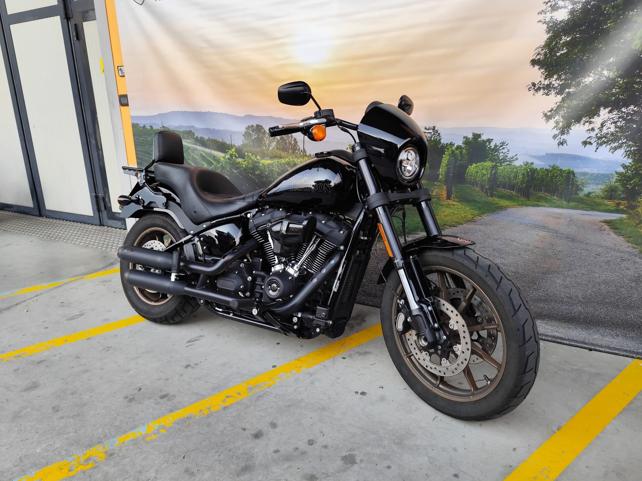 Harley-Davidson Low Rider S nera parcheggiata su asfalto con strisce gialle, sfondo con paesaggio collinare e vigneti al tramonto.