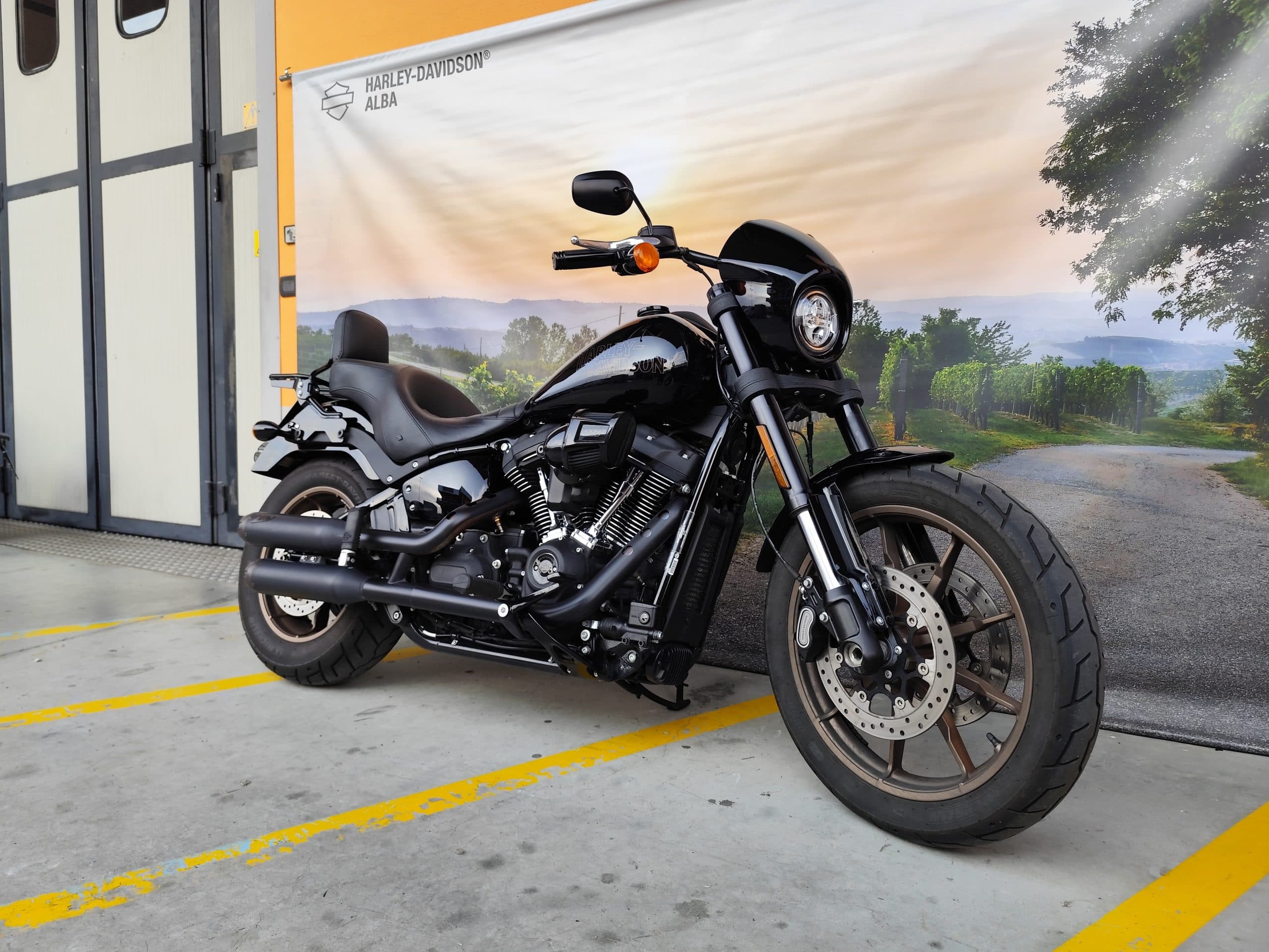 Moto Harley-Davidson nera, stile cruiser, parcheggiata davanti a un banner promozionale Harley-Davidson Alba con paesaggio collinare e vigneti.
