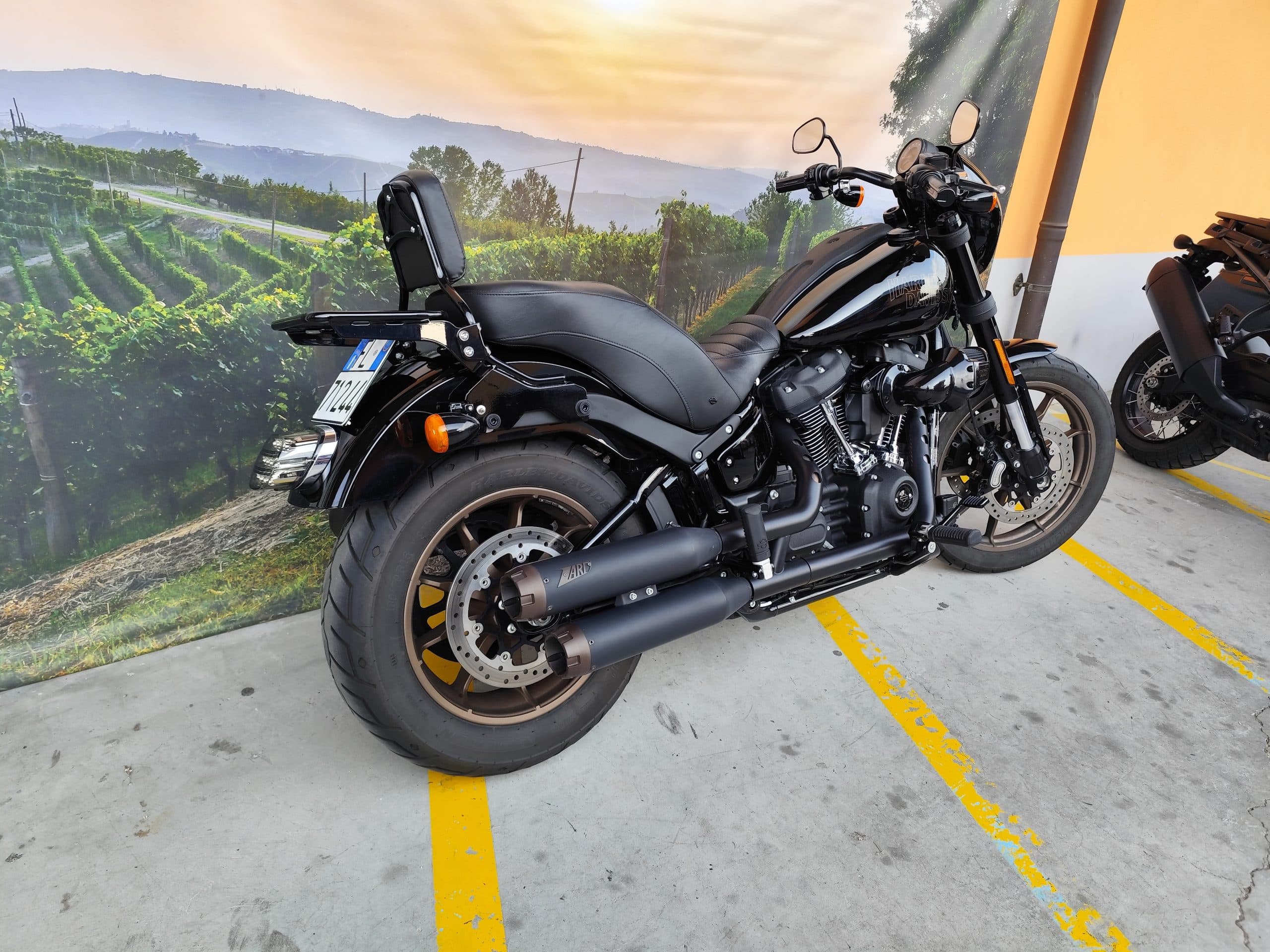 Harley-Davidson nera con cerchi bronzo e scarichi custom, parcheggiata su cemento. Fondale scenografico di vigneti al tramonto, suggerendo un test ride.