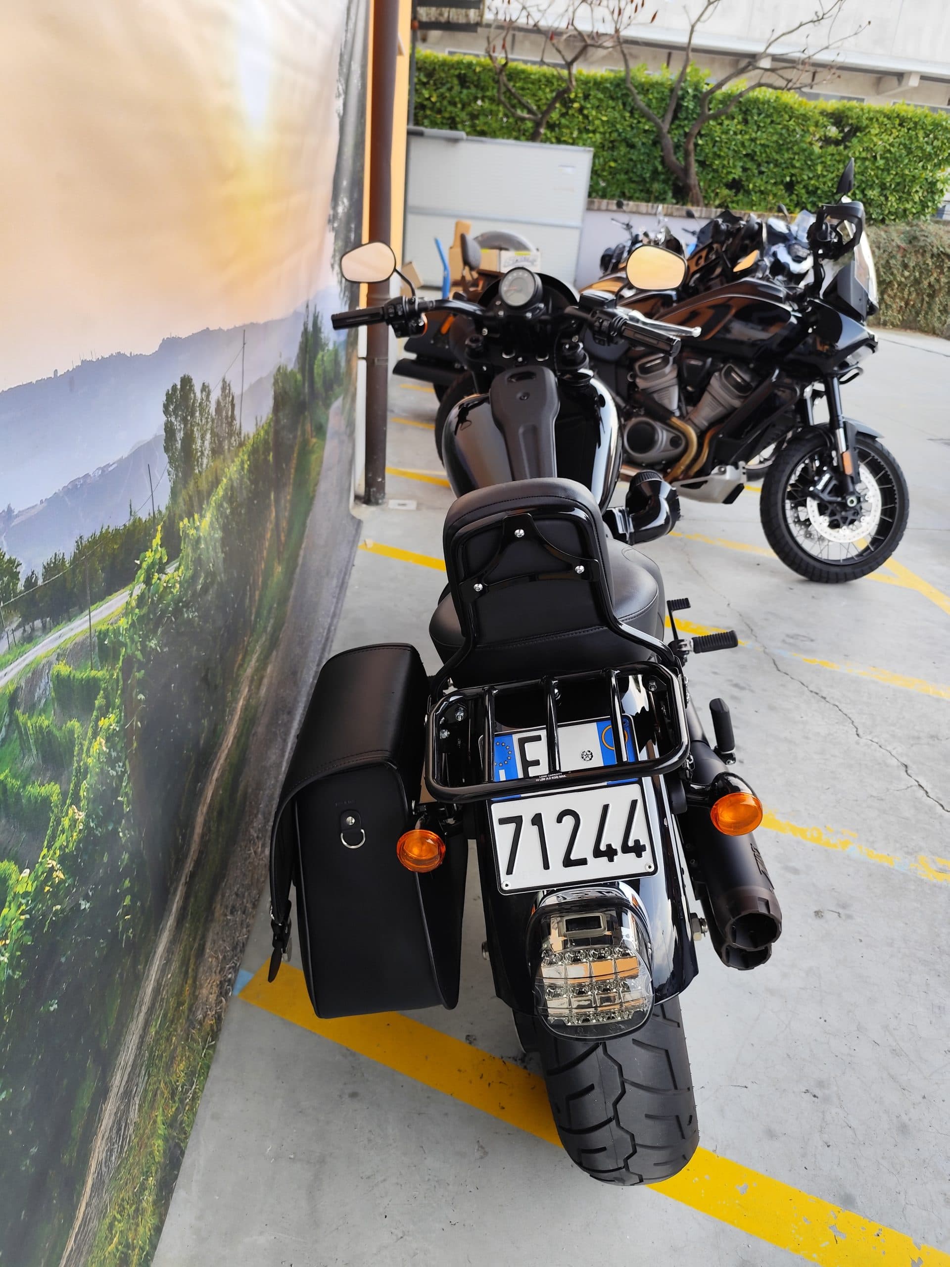 Harley-Davidson nera con borse laterali, targa F 71244, in area espositiva. Altre moto e murale paesagg