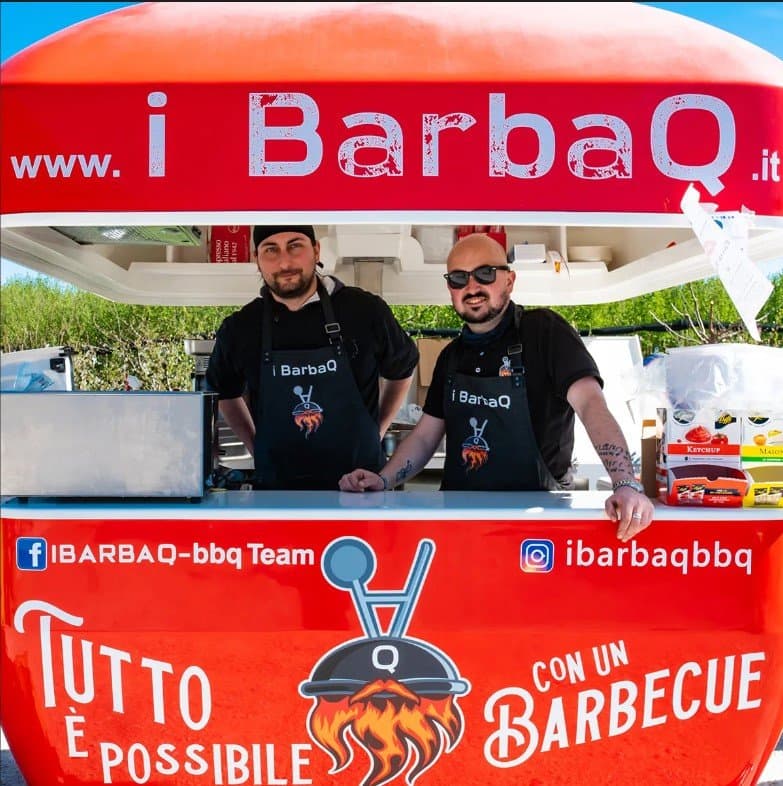 Due uomini sorridenti dietro un food truck rosso brandizzato 'i BarbaQ', specializzato in barbecue, con logo di grill e barba fiammante.