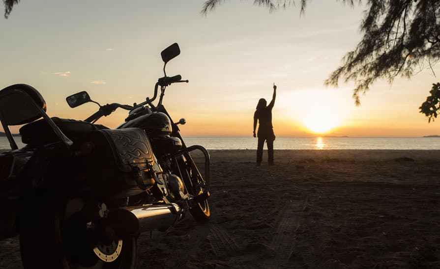 Harley-Davidson su spiaggia al tramonto, con motociclista in silhouette che alza un braccio verso l'orizzonte marino.