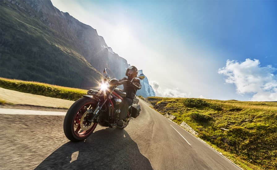 Moto Harley-Davidson in curva su strada alpina, pilota con abbigliamento tecnico. Faro acceso con bagliore solare. Ideale per test ride.