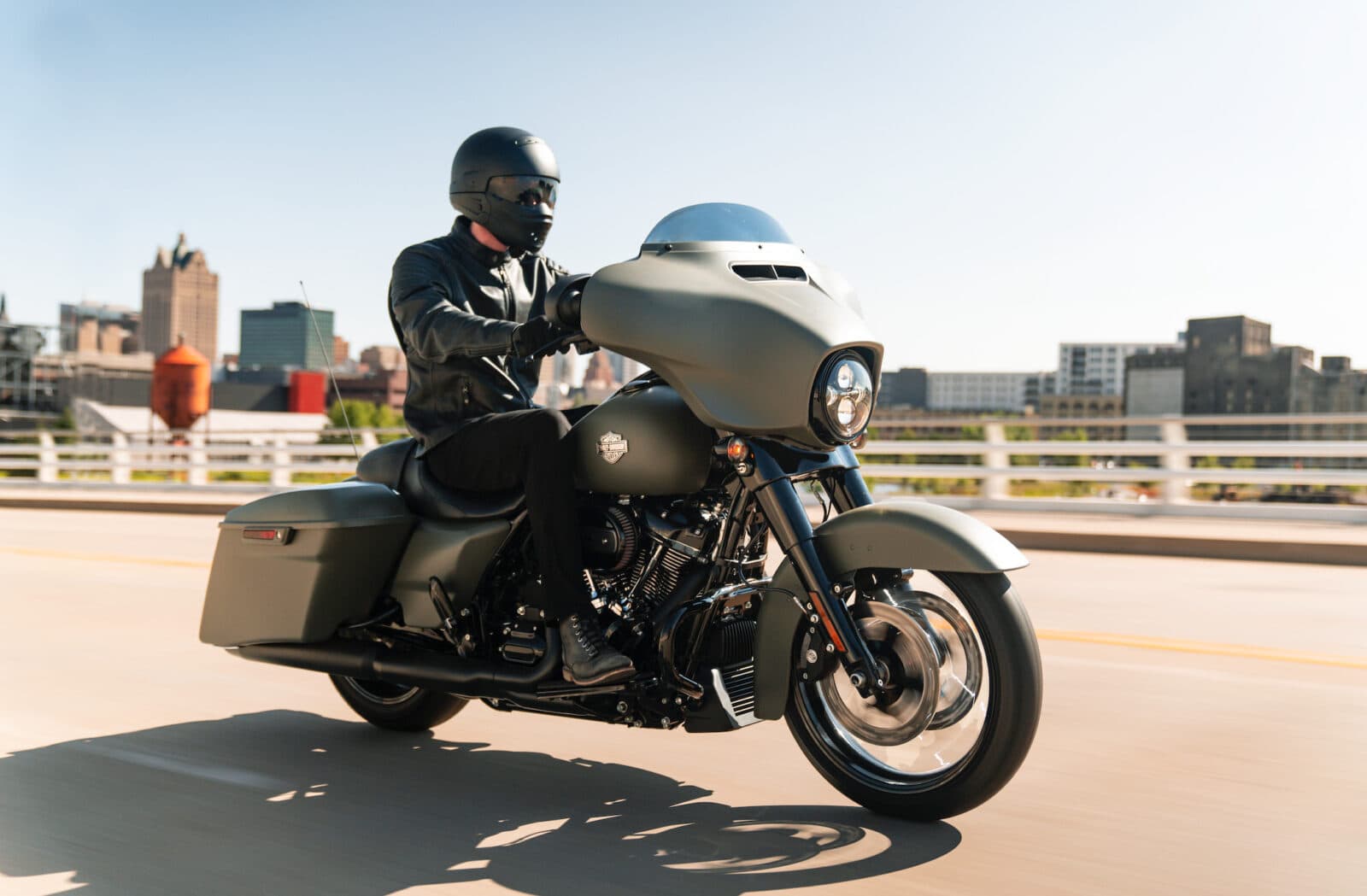Motociclista con abbigliamento tecnico su Harley-Davidson touring verde opaco in movimento su ponte. Skyline urbano. Perfetta per un test ride.