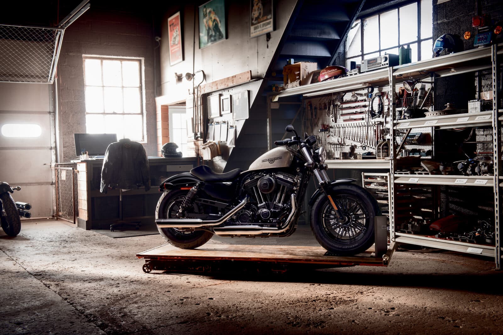 Harley-Davidson Sportster su pedana espositiva in officina, con giacca in pelle e casco su scrivania, attrezzi e ricambi.