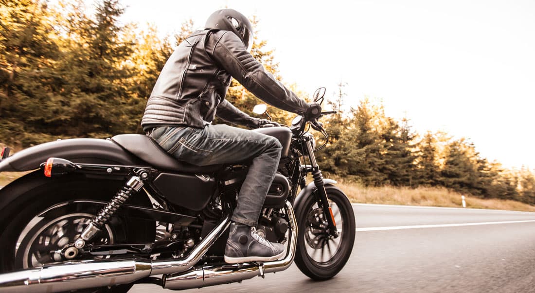 Motociclista con casco, giacca in pelle e abbigliamento tecnico su moto Harley-Davidson in movimento su strada, evocando l'emozione del test ride.