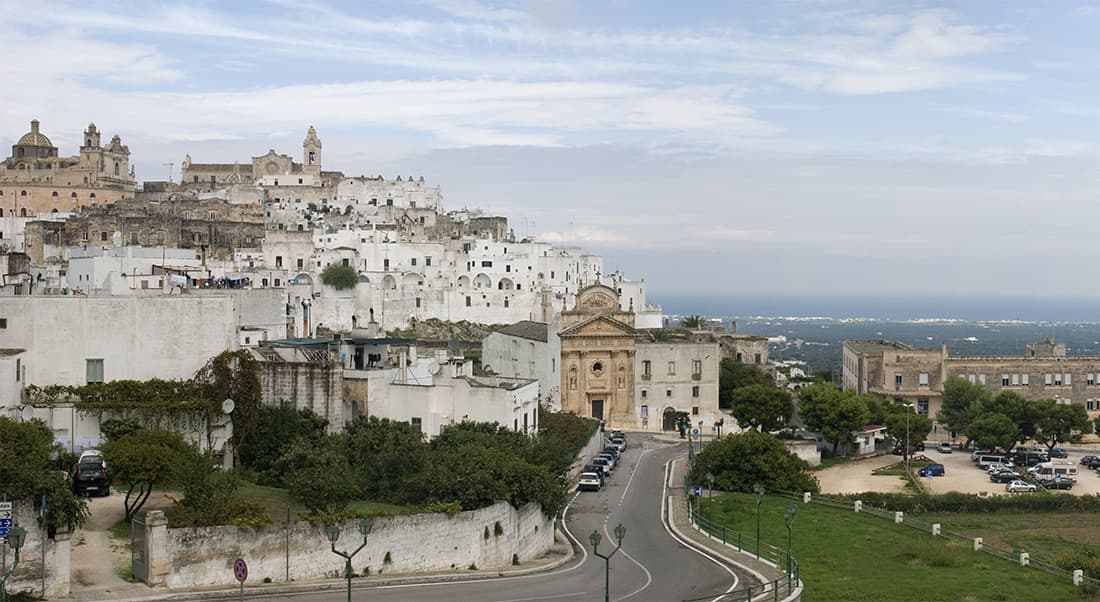 Vista panoramica di una città collinare mediterranea, caratterizzata da edifici bianchi e chiese storiche, con il mare in lontananza.