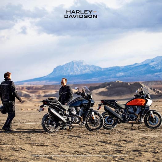 Due motociclisti con abbigliamento Harley-Davidson accanto a tre moto Pan America in un paesaggio montano desertico. Ideale per test ride e avventura.