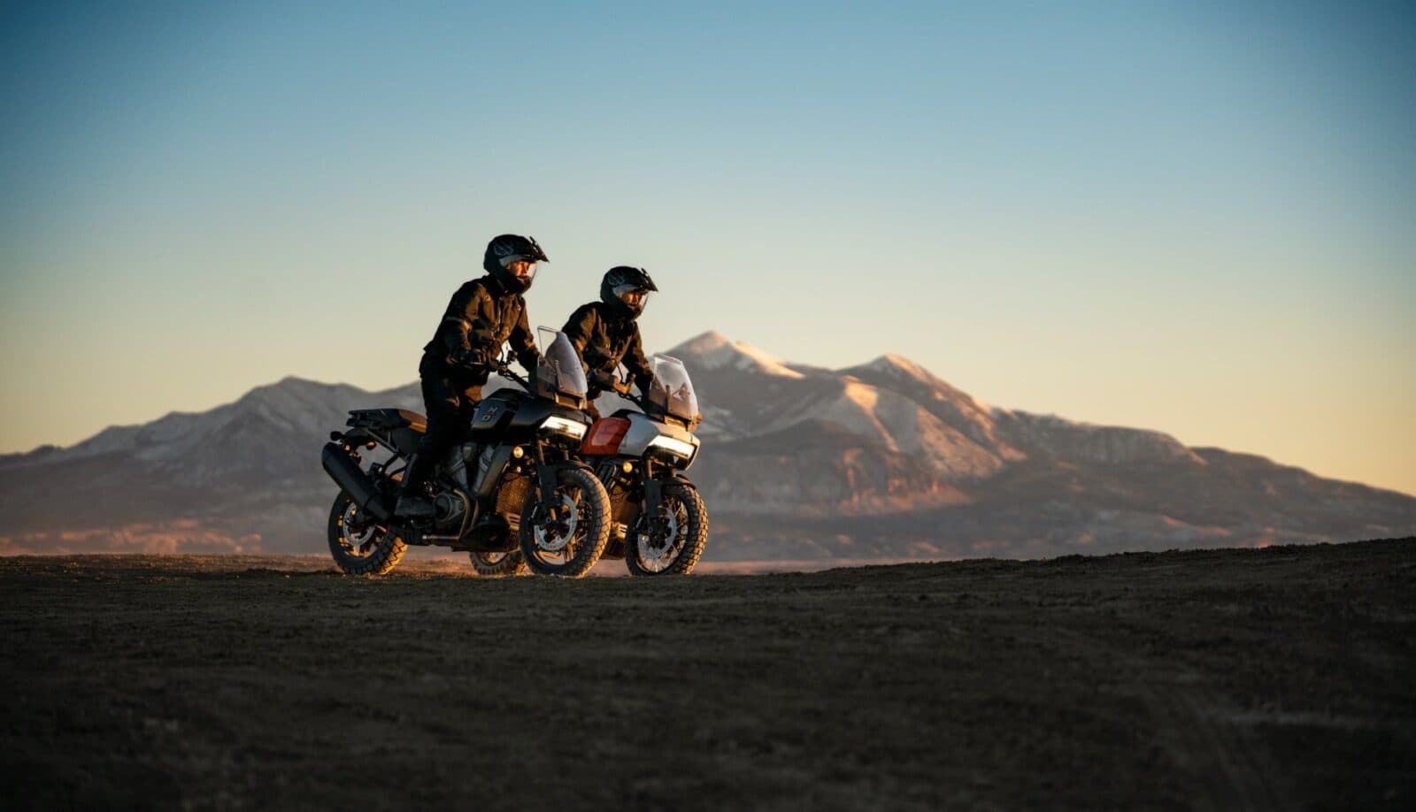 Due motociclisti con abbigliamento tecnico su moto Harley-Davidson Pan America in test ride off-road, con montagne innevate al tramonto.