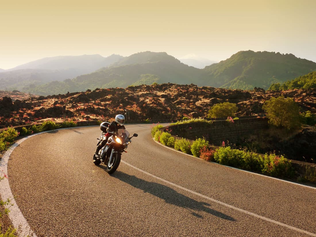 Pilota con abbigliamento tecnico e casco in sella a una moto su strada panoramica. Curva su asfalto tra rocce vulcaniche e colline.