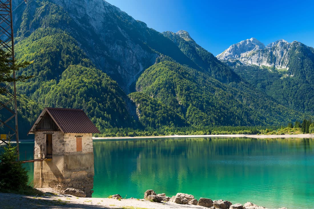 Paesaggio montano con lago alpino dalle acque turchesi e smeraldo, riflettente le cime boscose. In primo piano, una piccola casa in pietra parzialmente sommersa.