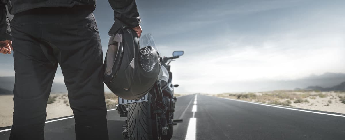 Motociclista in abbigliamento tecnico con casco, accanto alla sua moto, su una strada che si estende all'orizzonte. L'emozione
