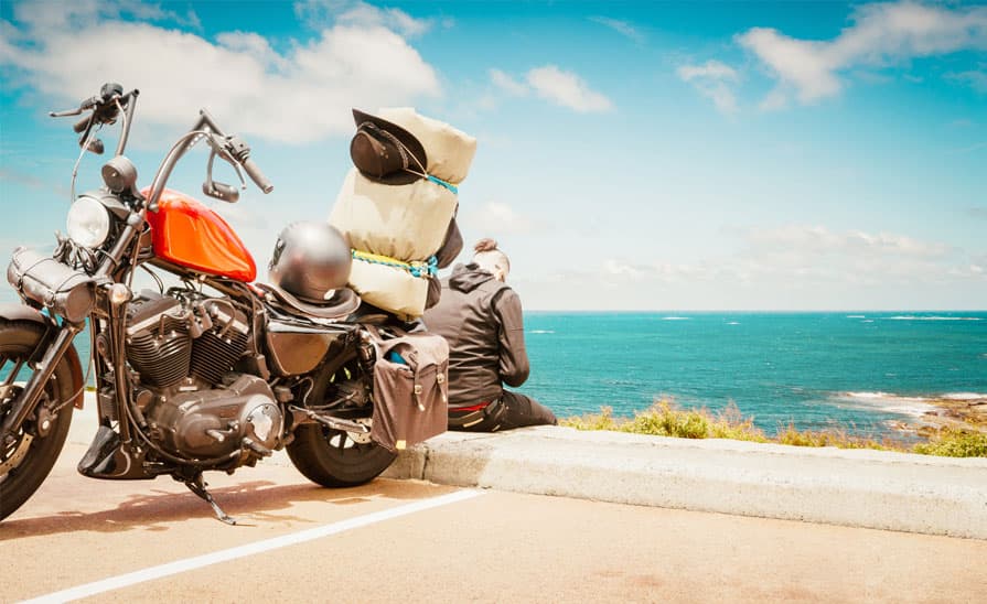 Harley-Davidson Sportster rossa con bagagli e casco, parcheggiata su strada panoramica. Un motociclista ammira il mare, simbolo di libertà e viaggio.
