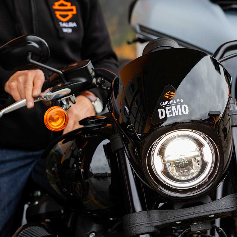 Primo piano di una moto Harley-Davidson nera con faro LED acceso e adesivo "GENUINE H-D DEMO" sul cupolino. Un uomo con felpa Harley-Davidson e orologio al polso afferra la leva del freno anteriore.