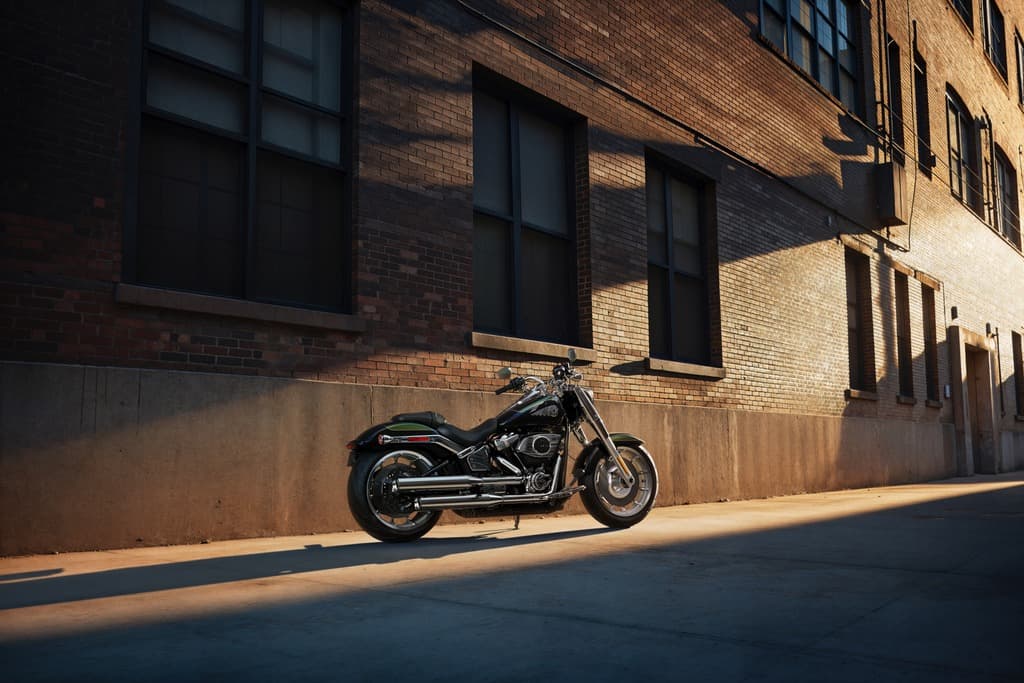Harley-Davidson nera parcheggiata su strada asfaltata, illuminata da luce radente, con edificio in mattoni e finestre scure sullo sfondo.