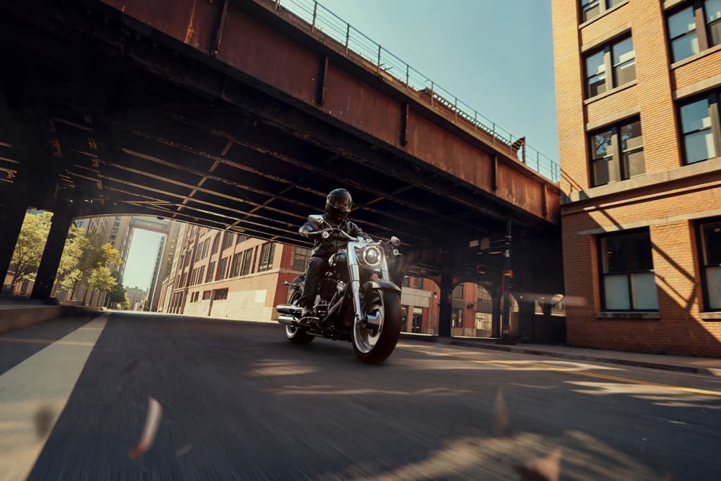 Motociclista con casco e giacca in pelle su moto Harley-Davidson in corsa su strada urbana, sotto un viadotto. Edifici in mattoni.