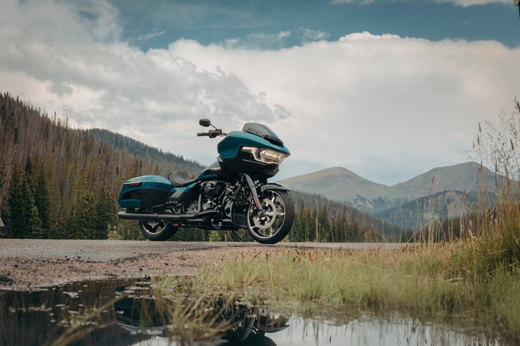 Harley-Davidson Road Glide Special blu-verde parcheggiata su strada di montagna, con fari accesi e riflesso in pozzanghera, tra foreste e cime.