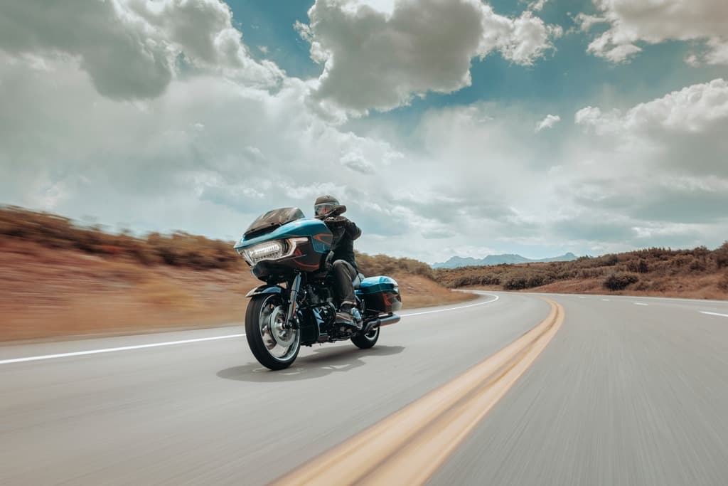 Motociclista con casco e abbigliamento tecnico in sella a una Harley-Davidson touring blu su strada panoramica con curve, cielo nuvoloso.