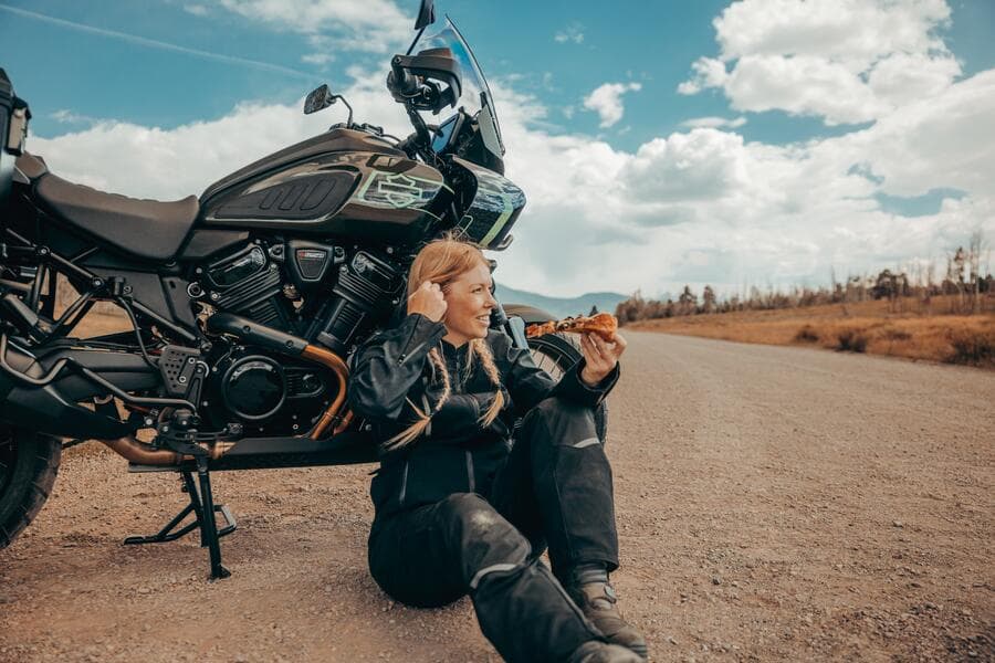Donna motociclista in abbigliamento tecnico Harley-Davidson Pan America, seduta a bordo strada, sorride mentre mangia pizza.