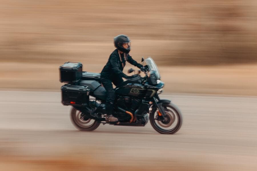 Motociclista con casco nero e abbigliamento tecnico scuro su una Harley-Davidson Pan America nera, dotata di bauletto e borse laterali rigide, in movimento su strada sterrata con effetto panning.