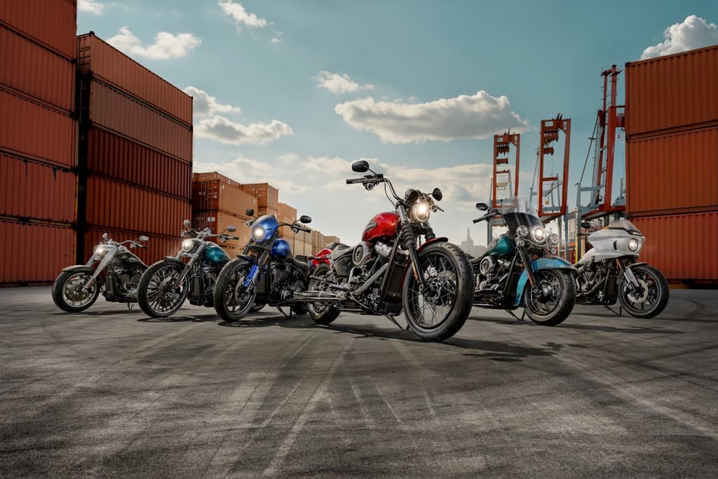 Sette motociclette Harley-Davidson di vari modelli e colori, parcheggiate in un'area portuale con container e gru. Evidenzia la gamma e la potenza dei motori.