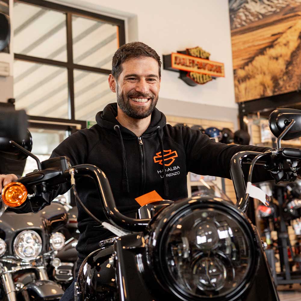Uomo sorridente con felpa Harley-Davidson Alba Lorenzo su moto H-D nera in concessionaria. Promuove abbigliamento e l'esperienza di test ride.