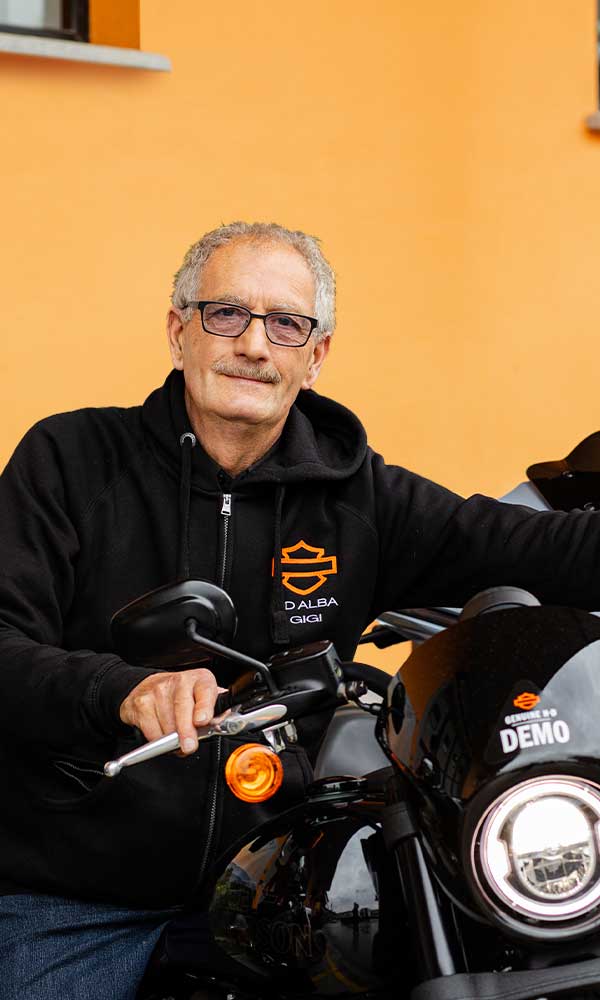 Uomo con felpa nera Harley-Davidson personalizzata 'DALBA GIGI' in sella a una moto Harley-Davidson demo nera, con adesivo 'GENUINE H-D DEMO' sul cupolino.