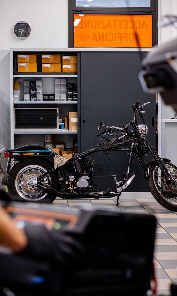 Officina autorizzata Harley-Davidson: moto in fase di lavorazione con telaio e motore a vista, affiancata da scaffali di accessori e ricambi originali.