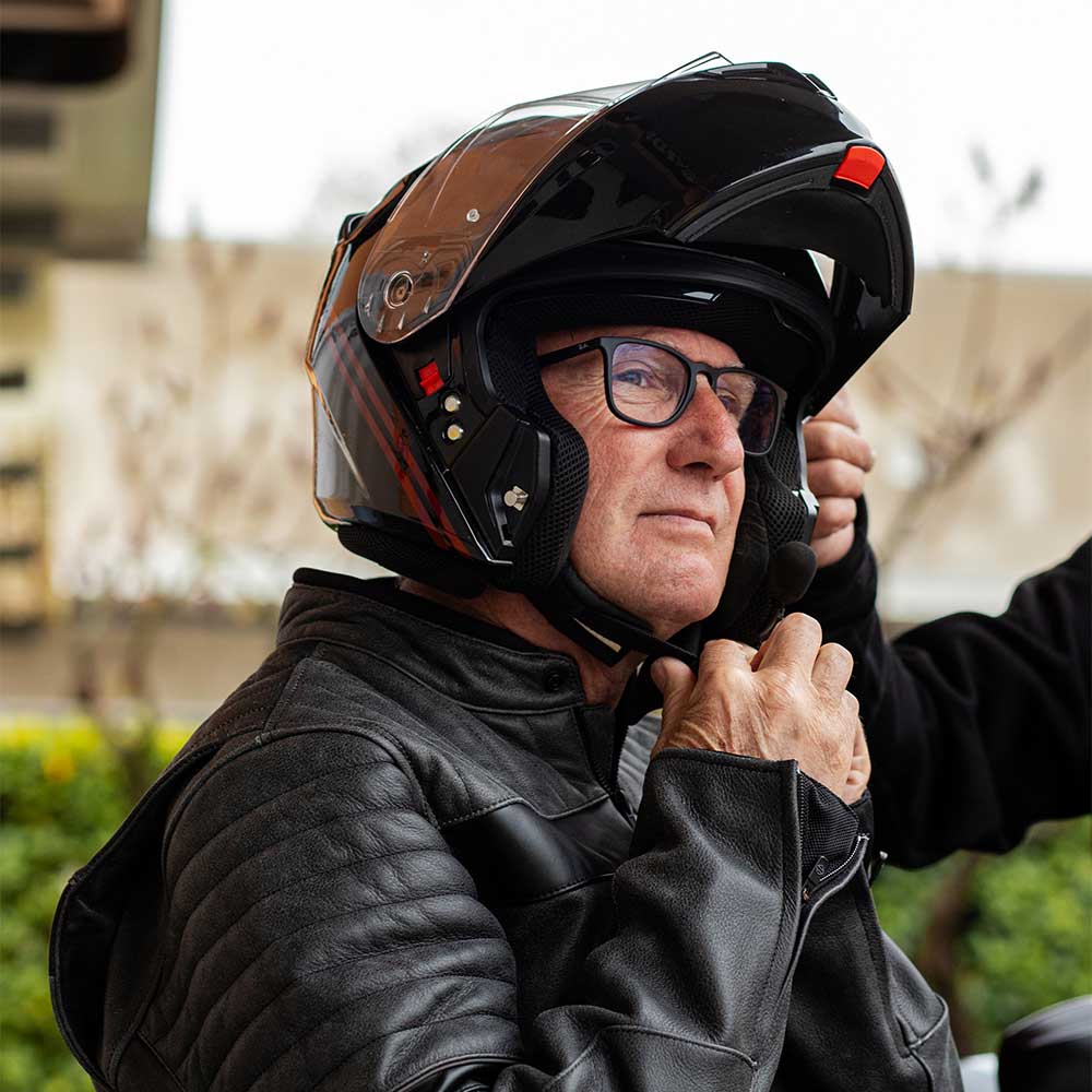 Uomo con casco modulare nero e giacca in pelle, regola il sottogola del casco con interfono, preparandosi per un test ride in moto.