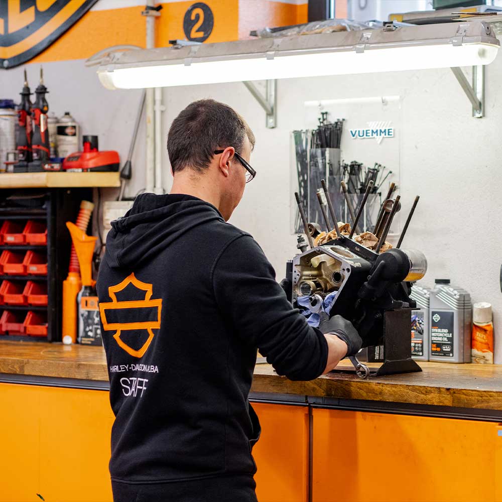 Tecnico Harley-Davidson di Alba con felpa brandizzata, assembla o pulisce un motore moto su banco officina, con attrezzi e oli specifici.