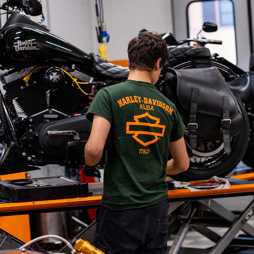 Tecnico Harley-Davidson di Alba in abbigliamento brandizzato, impegnato nella manutenzione di una moto nera in officina.