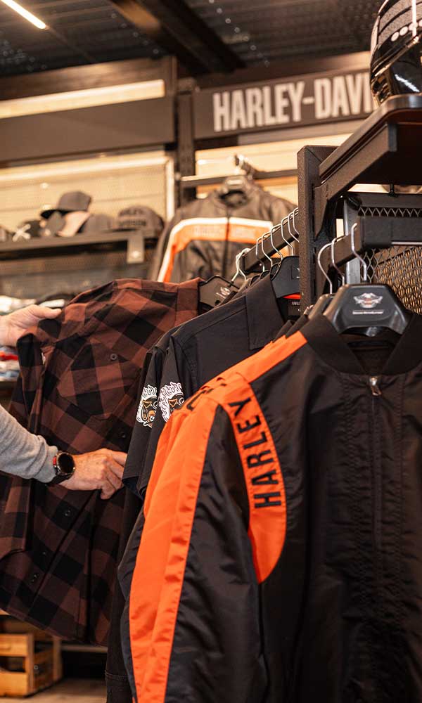 Cliente che esamina abbigliamento Harley-Davidson, tra cui una giacca nera con dettagli arancioni e scritta HARLEY, in una concessionaria.