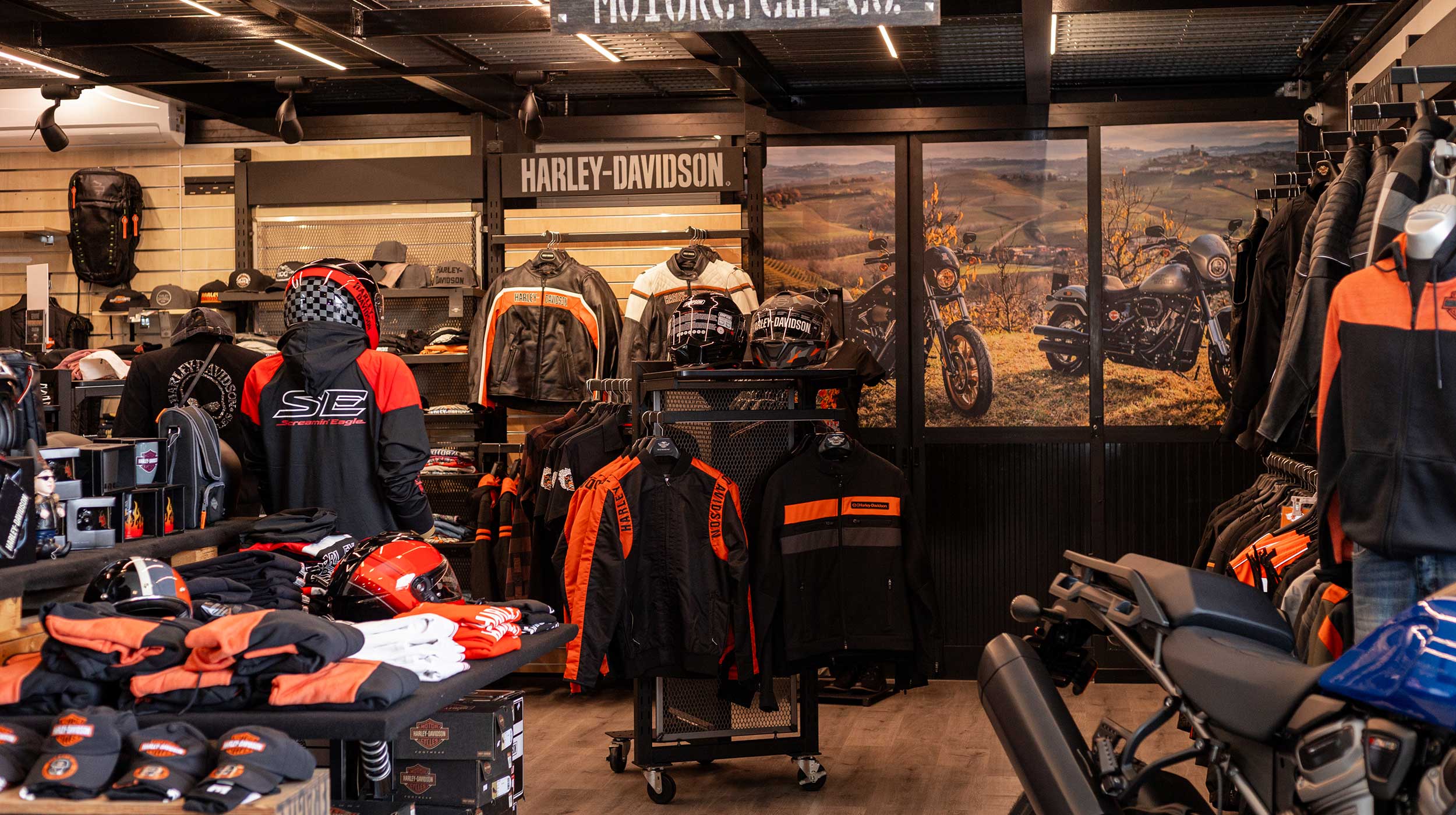 Concessionaria Harley-Davidson: esposizione di abbigliamento, caschi e accessori. Giacca Screamin' Eagle. Pannelli murali con moto e paesaggio.