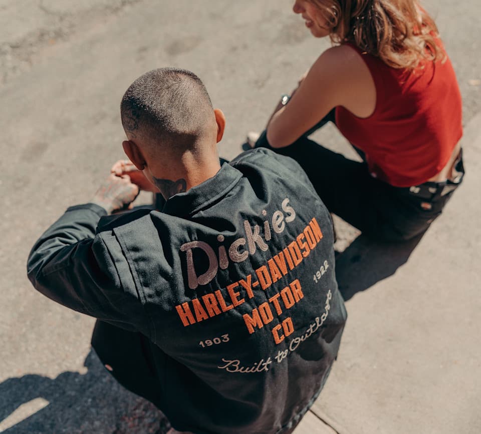 Schiena di persona con giacca nera Dickies Harley-Davidson ricamata, con loghi e date '1903' e '1922'. Ab