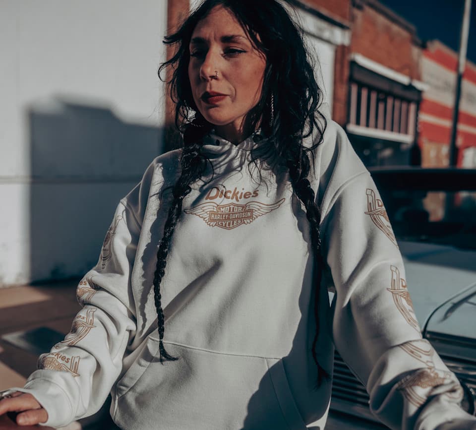 Modella indossa felpa con cappuccio Dickies Harley-Davidson bianca, con logo alato e grafiche distintive sulle maniche, in contesto urbano.