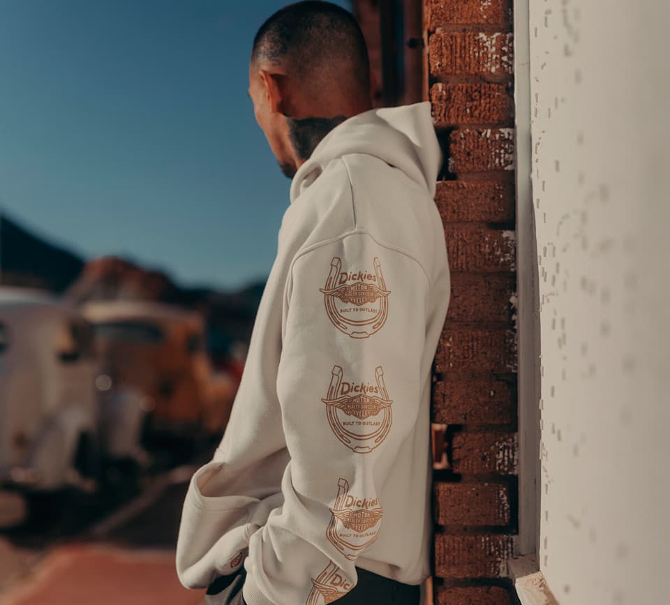 Uomo indossa felpa Dickies x Harley-Davidson con logo a ferro di cavallo e ali ripetuto sulla manica. Sfondo esterno sfocato.