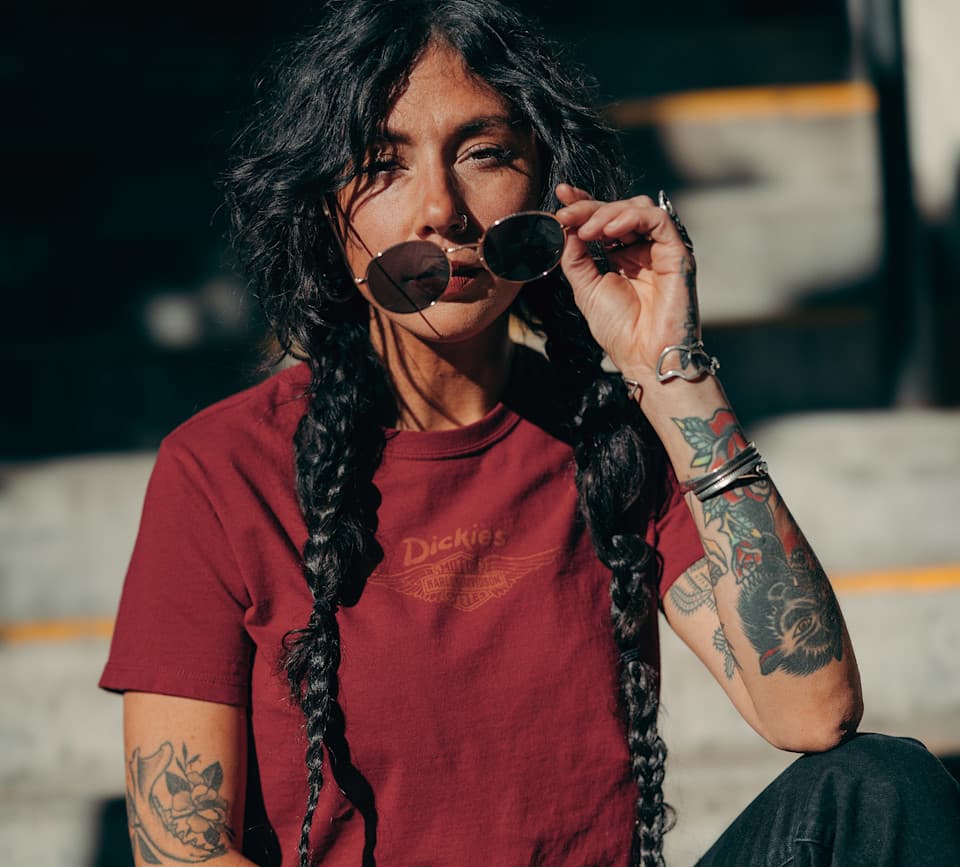 Ritratto di donna con trecce, indossa t-shirt Dickies con logo Harley-Davidson alato, occhiali da sole e tatuaggi. Espressione di stile H-D.