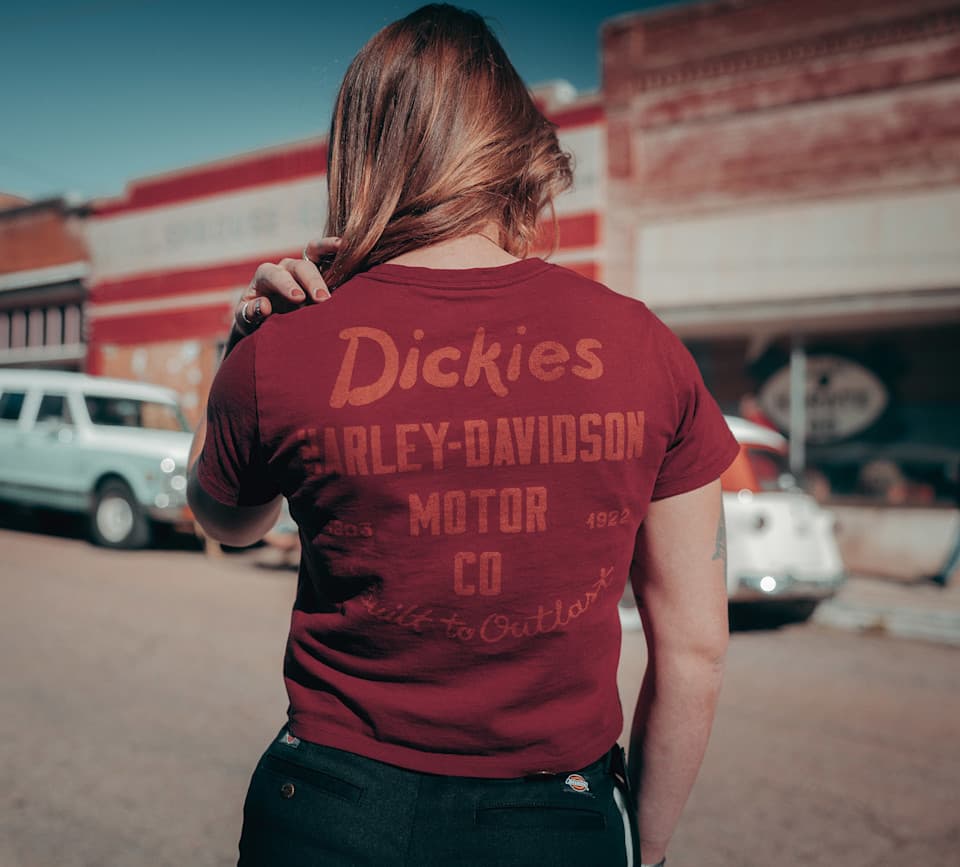 Persona di spalle con maglietta rossa Dickies e Harley-Davidson, con scritte 'MOTOR CO', '1903', '1922', 'Built to Outlaw'. Sfondo urbano con edifici e auto d'epoca.