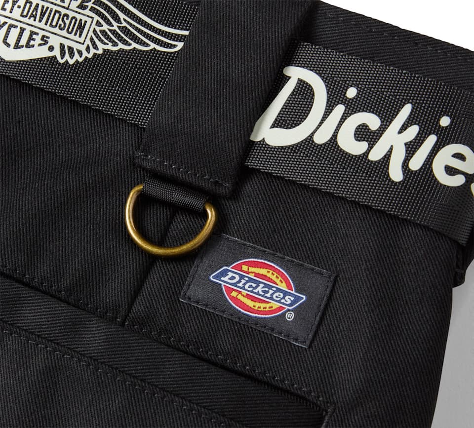 Primo piano di tessuto nero, probabilmente abbigliamento, con loghi Harley-Davidson e Dickies. Presenta un anello a D in ottone e una cinghia con