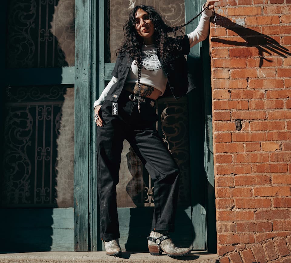 Donna con capelli ricci, indossa giacca nera, pantaloni Dickies neri e stivali effetto serpente. Appoggiata a porta blu, con tatuaggi e accessori.